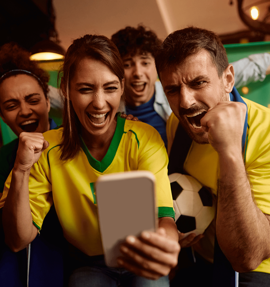 Four excited people in yellow and blue sports jerseys cheer while looking at a smartphone, with one holding a soccer ball.